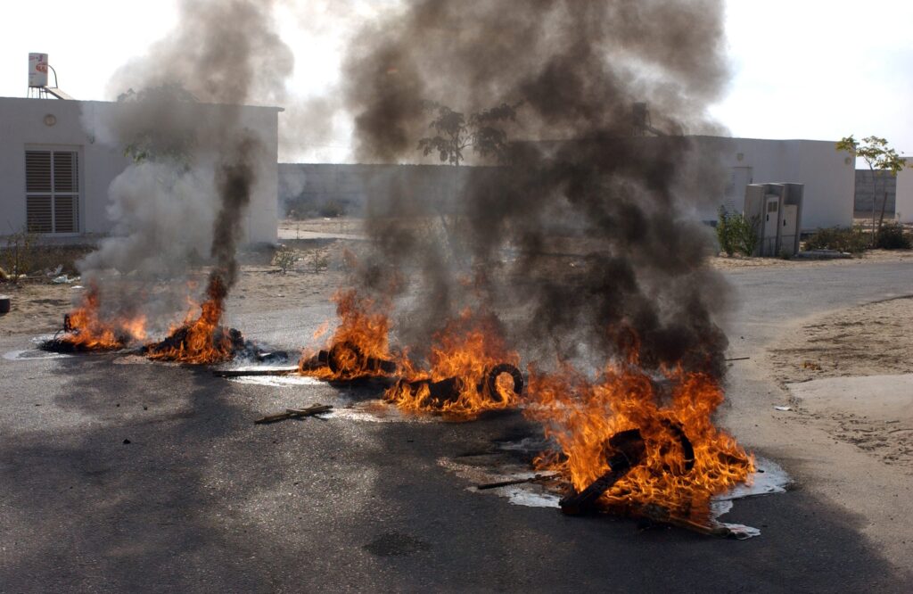 [בתמונה: צמיגים בוערים בכניסה לישוב מורג, למניעת כניסת כוחות הביטחון הבאים לפנות את תושבי המקום... מבט לאחור על אירועי קיץ 2005, אינו דומה להתכנסות זיכרון רגילה. הדיון שלפנינו טעון בשאיפות מעשיות לעתיד. ראוי לפיכך להכיר במלוא עומקה האקטואלי של המחלוקת ובהשקפות העולם השונות היוצרות אותה. קרדיט: MARK NEYMAN, לע"ם]
